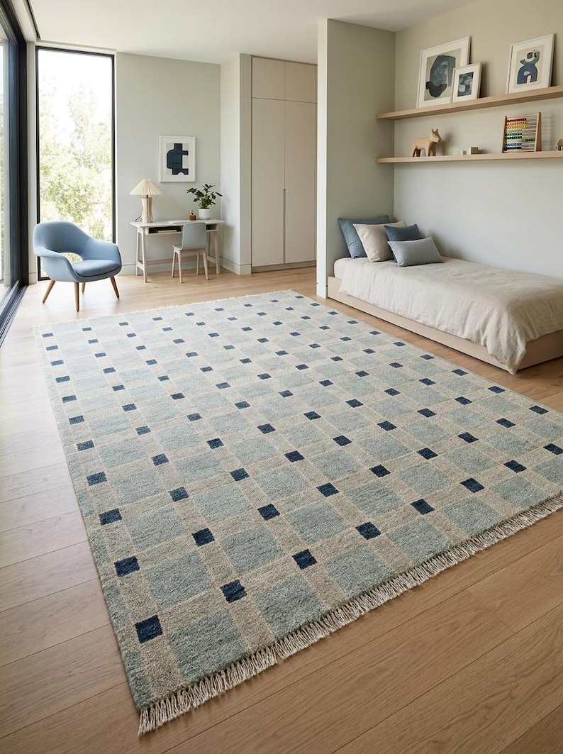 K&ouml;nnte beinhalten: Ein rechteckiger Teppich mit einem Gittermuster aus hellblauen und beigen Quadraten, wobei sich an jedem Schnittpunkt ein dunkelblaues Quadrat befindet. Der Teppich hat eine strukturierte Oberfl&auml;che und kurze wei&szlig;e Fransen an den kurzen Seiten.
