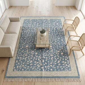 May include: A rectangular area rug featuring a dusty blue base with an off-white speckled pattern and a cream-colored Greek key border. The rug has short fringe detailing along the top and bottom edges, providing a textured finish to the design.