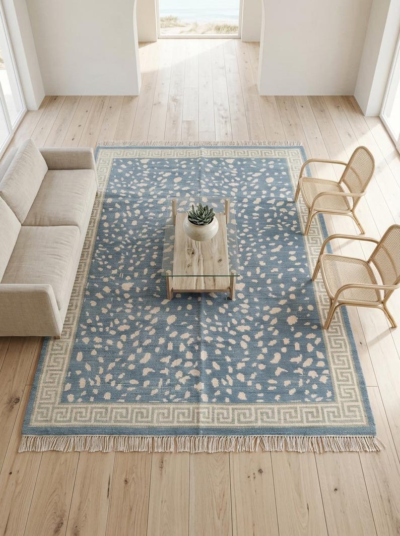 May include: A rectangular area rug featuring a dusty blue base with an off-white speckled pattern and a cream-colored Greek key border. The rug has short fringe detailing along the top and bottom edges, providing a textured finish to the design.