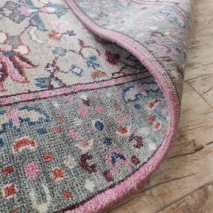 Könnte beinhalten: Ein gewebter Teppich mit einem detaillierten Blumen- und geometrischen Muster in Grau-, Rosa-, Blau- und Cremetönen. Der Teppich hat einen dicken, strukturierten Flor und einen markanten rosa Rand, wobei eine Ecke umgeschlagen ist.