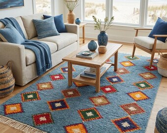 Puede incluir: Una alfombra rectangular con una base de color azul claro texturizada y un patrón repetitivo de diamantes multicolores en rojo, naranja, amarillo y verde. La alfombra está terminada con flecos de color crema en los dos bordes más cortos.