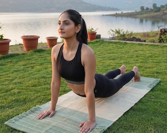 May include: A rectangular yoga mat featuring a two-tone design with a sage green section on one end and a cream-colored section on the other. The mat has a textured woven surface and decorative fringe detailing along the short edges.