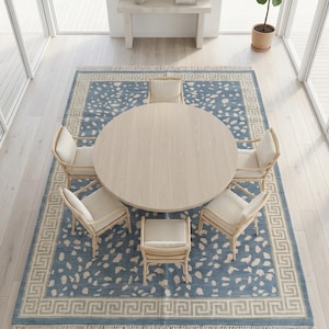May include: A round light wood dining table surrounded by five chairs with light-colored cushions, placed on a large rectangular blue rug featuring a cream-colored speckled pattern and a Greek key border with fringe on the short ends.