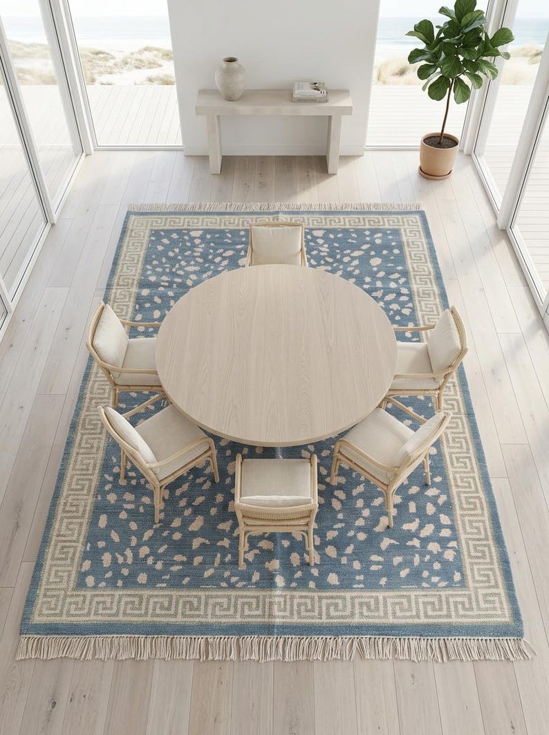 May include: A round light wood dining table surrounded by five chairs with light-colored cushions, placed on a large rectangular blue rug featuring a cream-colored speckled pattern and a Greek key border with fringe on the short ends.