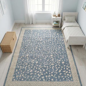May include: A rectangular area rug featuring a dusty blue base with an all-over pattern of irregular cream-colored spots. The rug is framed by a wide border with a repeating geometric Greek key design and finished with short fringe on both ends.