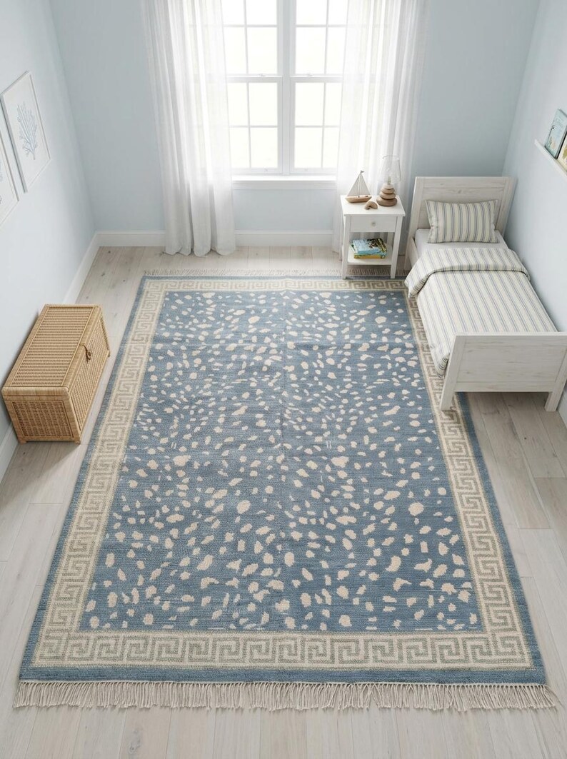 May include: A rectangular area rug featuring a dusty blue base with an all-over pattern of irregular cream-colored spots. The rug is framed by a wide border with a repeating geometric Greek key design and finished with short fringe on both ends.