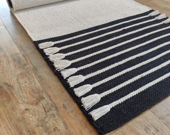 May include: A rectangular woven rug featuring a solid off-white section at one end and a black section with white horizontal stripes at the other. The black area is accented with white fringe tassels attached to the ends of the white stripes.