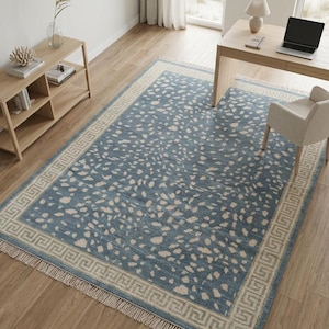 May include: A rectangular area rug featuring a dusty blue base with an irregular cream-colored speckled pattern. The rug is framed by a wide border with a geometric Greek key design and finished with short, light-colored fringe on both ends.