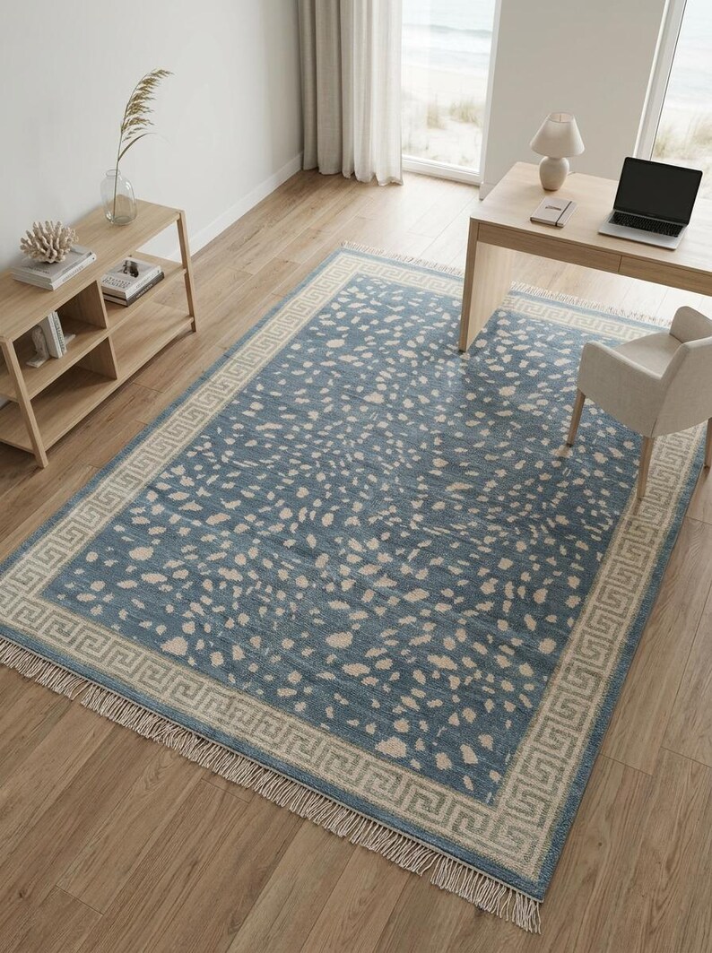 May include: A rectangular area rug featuring a dusty blue base with an irregular cream-colored speckled pattern. The rug is framed by a wide border with a geometric Greek key design and finished with short, light-colored fringe on both ends.