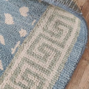 May include: A light blue and cream area rug featuring a textured pile surface. The rug displays a central pattern of small, irregular cream-colored shapes on a light blue background, bordered by a distinct geometric Greek key design in cream.