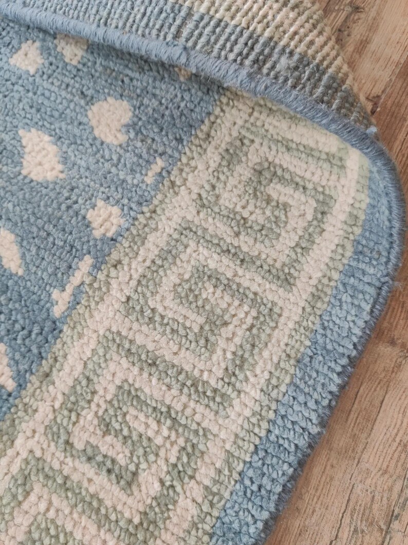 May include: A light blue and cream area rug featuring a textured pile surface. The rug displays a central pattern of small, irregular cream-colored shapes on a light blue background, bordered by a distinct geometric Greek key design in cream.