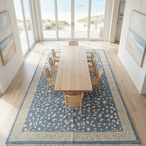 May include: A rectangular area rug featuring a blue field with an off-white abstract spotted pattern and a Greek key border design. The rug has a fringe trim on both ends and is positioned under a long light-colored wooden dining table with chairs.