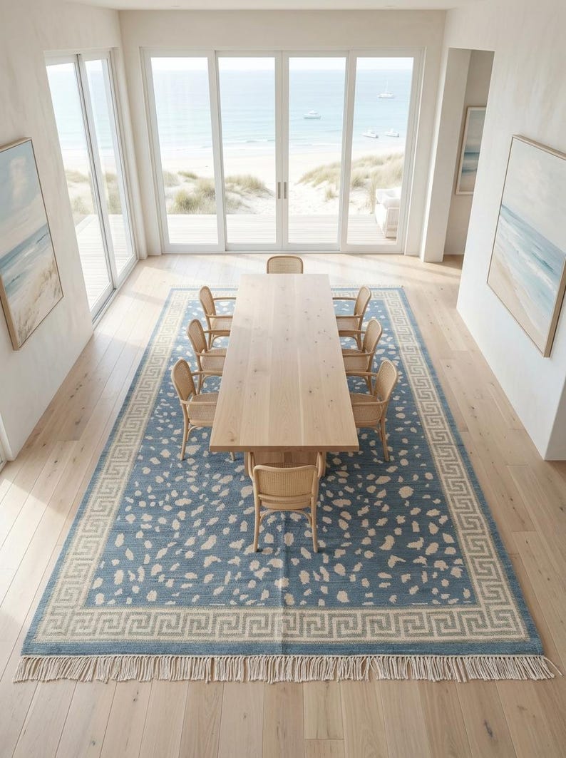 May include: A rectangular area rug featuring a blue field with an off-white abstract spotted pattern and a Greek key border design. The rug has a fringe trim on both ends and is positioned under a long light-colored wooden dining table with chairs.