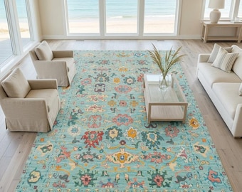 May include: A large rectangular area rug featuring a light blue background with a repeating pattern of stylized floral and geometric motifs in shades of pink, yellow, dark blue, and cream, finished with a short white fringe on the ends.