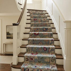 May include: A staircase featuring a blue floral runner rug secured with brass stair rods. The stairs have dark wood treads and white risers, with a curved wooden handrail and white balusters leading up to a landing in a bright, neutral-toned hallway.