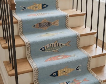 May include: A light blue stair runner rug featuring a repeating pattern of stylized fish in various colors including yellow, orange, and blue. The runner has a decorative cream-colored border with a geometric pattern and fringe at the bottom edge.