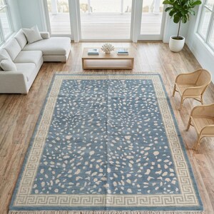 May include: A large rectangular area rug featuring a dusty blue field with an all-over cream-colored speckled pattern. The rug is framed by a wide cream border with a repeating geometric Greek key motif and finished with short fringe on both ends.