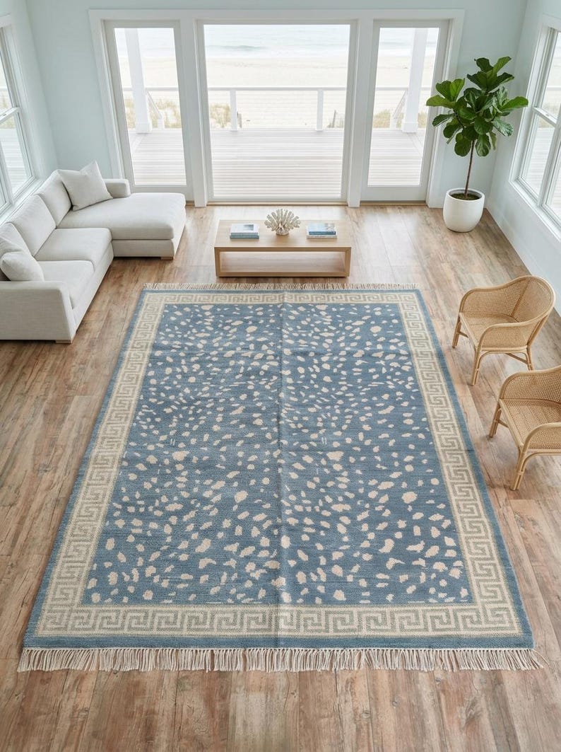 May include: A large rectangular area rug featuring a dusty blue field with an all-over cream-colored speckled pattern. The rug is framed by a wide cream border with a repeating geometric Greek key motif and finished with short fringe on both ends.