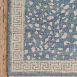 May include: A rectangular area rug featuring a blue field with an irregular cream-colored spotted pattern. The rug is framed by a wide border with a repeating geometric Greek key design in cream and light green, finished with a cream fringe.