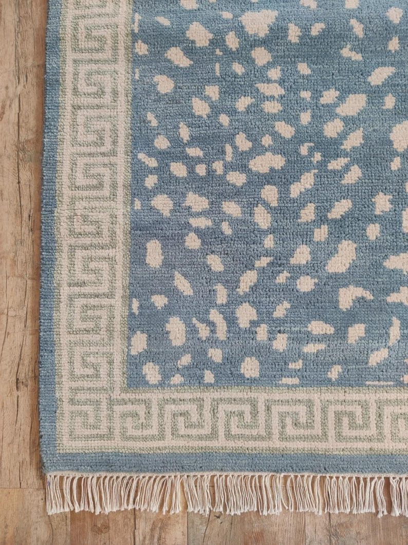 May include: A rectangular area rug featuring a blue field with an irregular cream-colored spotted pattern. The rug is framed by a wide border with a repeating geometric Greek key design in cream and light green, finished with a cream fringe.
