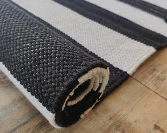 May include: A rectangular woven rug featuring a bold pattern of alternating black and white stripes. The rug is partially rolled up, revealing a thick, textured weave and a consistent striped design that continues across the entire surface area.
