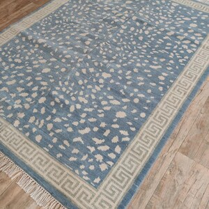May include: A rectangular area rug featuring a dusty blue field with an all-over pattern of irregular off-white spots. The rug is framed by a wide border with a geometric Greek key design in off-white and blue, finished with off-white fringe.