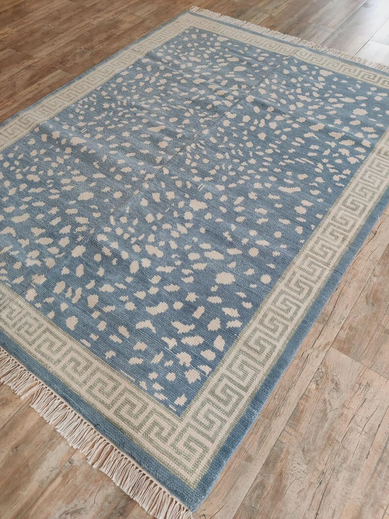 May include: A rectangular area rug featuring a dusty blue field with an all-over pattern of irregular off-white spots. The rug is framed by a wide border with a geometric Greek key design in off-white and blue, finished with off-white fringe.