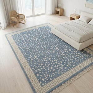 May include: A rectangular area rug featuring a dusty blue field with an all-over pattern of irregular cream-colored spots. The rug is framed by a wide border displaying a repeating geometric Greek key design in cream and blue, finished with fringe.