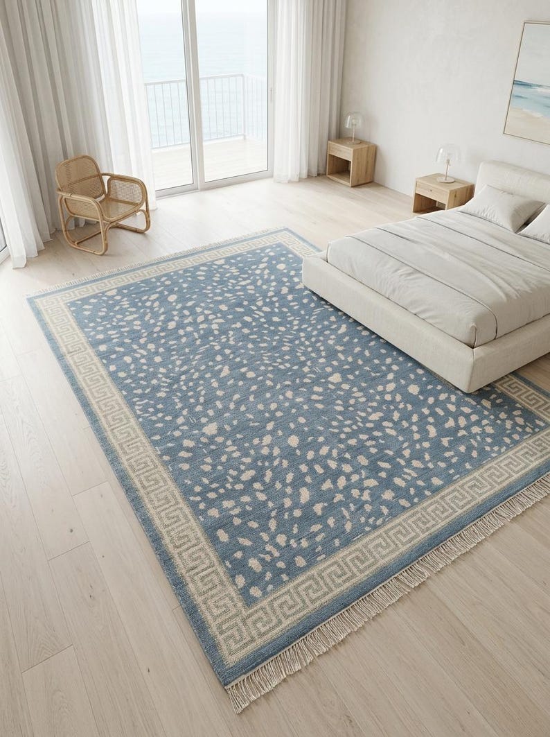 May include: A rectangular area rug featuring a dusty blue field with an all-over pattern of irregular cream-colored spots. The rug is framed by a wide border displaying a repeating geometric Greek key design in cream and blue, finished with fringe.