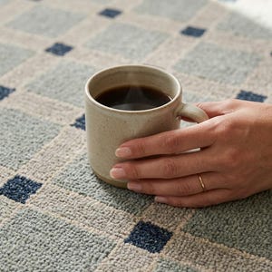 Peut inclure: Une tasse en céramique beige mouchetée remplie de café noir, posée sur un tapis texturé présentant un motif quadrillé de carrés bleu clair et de petits carrés bleu foncé aux intersections des lignes, créant un design géométrique.