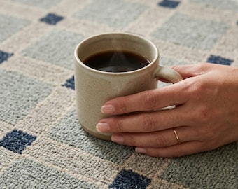 May include: A speckled beige ceramic mug filled with dark coffee, resting on a textured rug featuring a grid pattern of light blue squares and small dark blue squares at the intersections of the grid lines, creating a geometric design.
