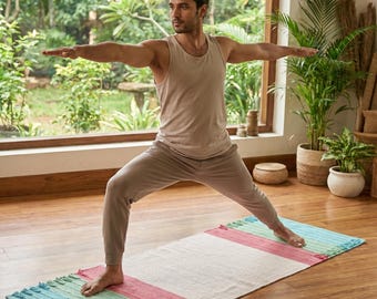 May include: A rectangular yoga mat featuring a woven texture with horizontal color blocks in light blue, mint green, and dusty rose, finished with short fringe detailing on both ends, positioned flat on a wooden floor surface.
