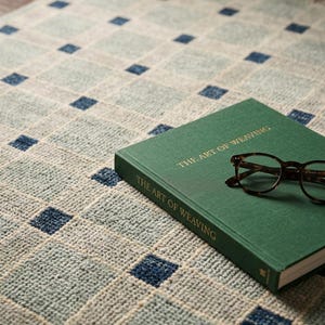 K&ouml;nnte beinhalten: Ein gewebter Teppich mit einem hellblauen und wei&szlig;en Gittermuster und dunkelblauen Quadraten an den Schnittpunkten. Ein gr&uuml;nes gebundenes Buch mit dem Titel THE ART OF WEAVING liegt auf dem Teppich, darauf eine braune Schildpattbrille.