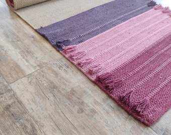 May include: A rectangular woven runner rug featuring a color-blocked design with sections of beige, deep purple, light pink, and dark magenta. The rug has a textured weave and is finished with decorative fringe tassels along the edges of the sections.