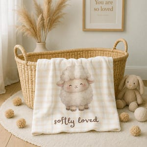 Könnte beinhalten: Eine cremefarbene und beige Decke mit Vichy-Muster, die in der Mitte eine Illustration eines flauschigen Schafs mit lächelndem Gesichtsausdruck zeigt. Unter dem Schaf ist der Text "softly loved" in brauner, abgerundeter Schreibschrift gedruckt.