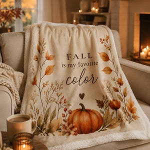 Könnte beinhalten: Eine cremefarbene Fleecedecke mit einem Aufdruck aus Herbstblättern, Zweigen und einem großen orangefarbenen Kürbis. Der Text "FALL is my favorite color" ist in schwarzer Schrift zu sehen, ergänzt durch ein kleines Herzsymbol über dem Kürbis.