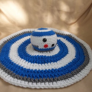 Puede incluir: Una manta de beb&eacute; de ganchillo con un patr&oacute;n de rayas azules, blancas y grises. Un juguete R2-D2 de ganchillo est&aacute; en el centro de la manta.