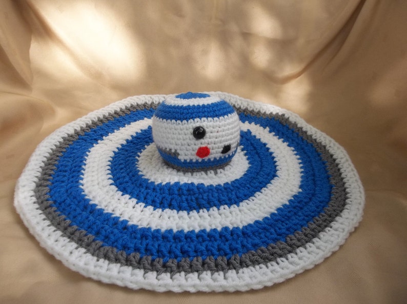 Puede incluir: Una manta de beb&eacute; de ganchillo con un patr&oacute;n de rayas azules, blancas y grises. Un juguete R2-D2 de ganchillo est&aacute; en el centro de la manta.