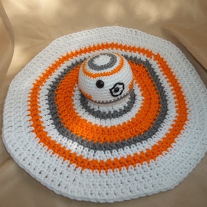 Puede incluir: Una alfombra redonda de crochet blanco con rayas naranjas y grises. Un peque&ntilde;o droide BB-8 de crochet est&aacute; en el centro de la alfombra.