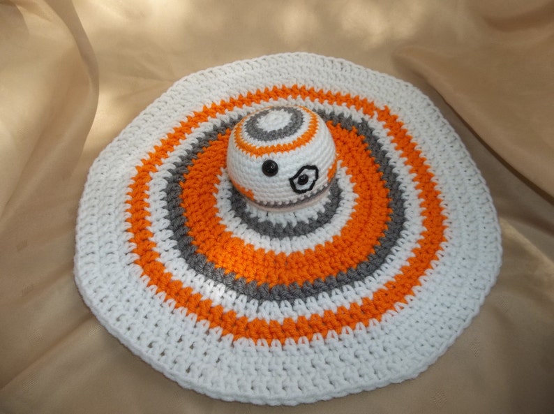 Puede incluir: Una alfombra redonda de crochet blanco con rayas naranjas y grises. Un peque&ntilde;o droide BB-8 de crochet est&aacute; en el centro de la alfombra.