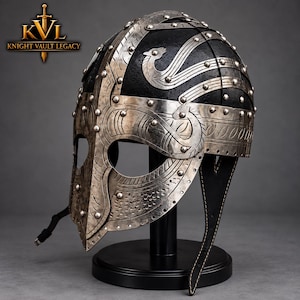 May include: A black metal helmet featuring silver-toned metal overlays with intricate engraved patterns and rivets. The helmet includes a nose guard, eye openings, and a leather strap. The text KVL KNIGHT VAULT LEGACY appears in the top left corner.
