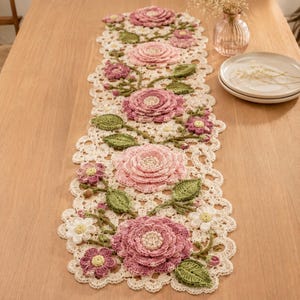 May include: A long, rectangular crochet table runner featuring an intricate cream-colored lace base adorned with three-dimensional floral motifs in shades of dusty pink, mauve, and white, accented by textured green crocheted leaves and stems.