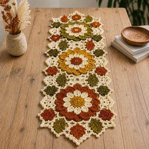 May include: A rectangular crochet table runner featuring a repeating floral and leaf pattern in shades of cream, olive green, mustard yellow, and burnt orange. The design consists of large central flower motifs connected by smaller flowers and leaves.