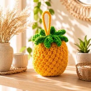May include: A crocheted pineapple-shaped object featuring a textured yellow body, a green leafy top, and a yellow loop handle extending from the center. The yarn construction creates a distinct, repeating pattern across the surface of the fruit.