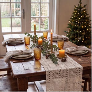 May include: A rustic wooden dining table set for a holiday meal with a cream-colored crocheted table runner, beige linen napkins, speckled ceramic plates, amber-colored textured glass tumblers, brass candle holders, and pillar candles.
