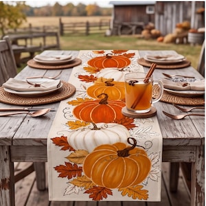 May include: A rectangular table runner featuring a printed pattern of orange and white pumpkins interspersed with brown, orange, and yellow autumn leaves on a cream-coloured fabric background, displayed centred on a rustic wooden dining table.