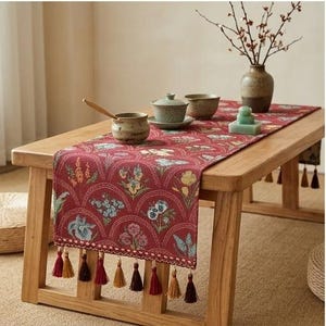Puede incluir: Un camino de mesa rectangular de color rojo con un patrón repetitivo de motivos florales dentro de segmentos arqueados. El camino de mesa está rematado con un fleco decorativo de pequeñas borlas multicolores en el borde.