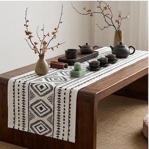Op de afbeelding: Een witte tafelloper met een zwart geometrisch patroon van ruiten en verticale strepen. De loper ligt op een houten tafel met een donkerbruine theepot, diverse kleine theekopjes, een keramische vaas met takken en een groen stenen object.