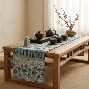 Puede incluir: Una mesa de té de madera con un camino de mesa azul claro con un patrón de árboles y flores de color marrón oscuro. Sobre la mesa hay una tetera marrón oscuro, un cuenco con tapa, tres tazas pequeñas sobre platillos de madera y un jarrón.