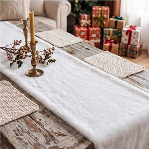 May include: A white faux fur table runner laid across a rustic wooden table, accompanied by two brass candle holders with yellow taper candles, a sprig of artificial berries, and three beige knitted placemats arranged on the surface.